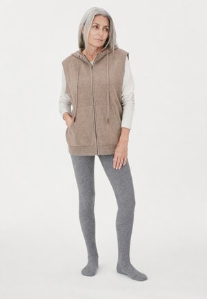 Femme âgée portant un sweat à capuche zippé sans manches beige, une chemise blanche à manches longues et des leggings gris, debout pieds nus sur un fond uni.