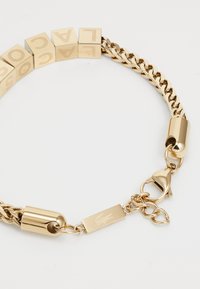 Bracelet en chaîne dorée avec des blocs carrés gravés de lettres, fermeture à fermoir en lobster et étiquette de logo, mettant en valeur une texture et un design métallique élégants.