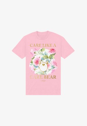 Růžové bavlněné tričko s květinovým designem, na kterém je medvěd a nápis "PEČUJ JAKO PÉČOVACÍ MEDVĚD 1982" ve zlaté barvě. Krátké rukávy, volný střih.