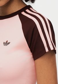 Krótki rękaw sportowego topu w jasnoróżowym i brązowym kolorze, z trzema brązowymi paskami na rękawach oraz haftowanym logo z przodu.