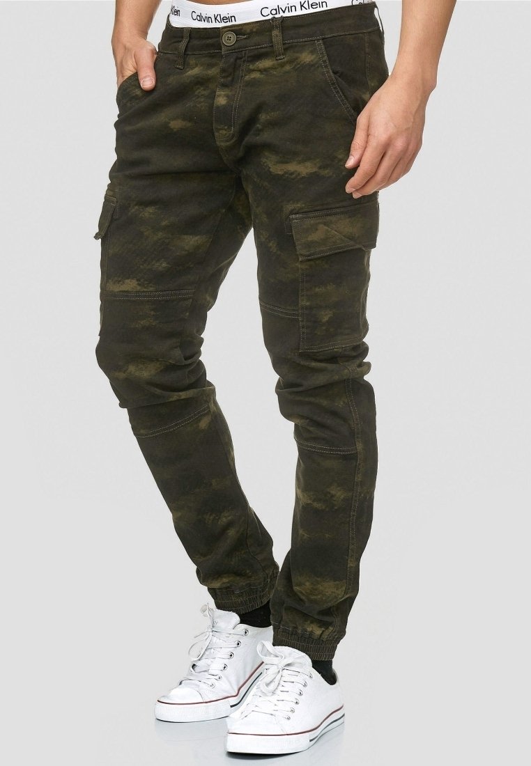 Homme portant un pantalon cargo camouflage vert foncé avec des poignets élastiques et des baskets blanches, mains dans les poches, debout devant un fond gris.