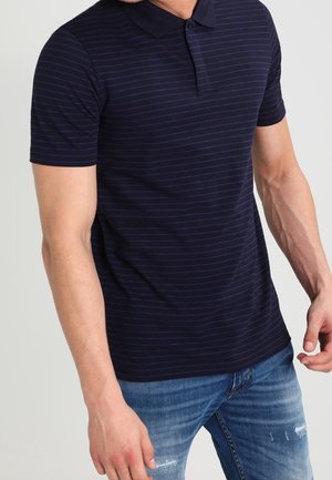 Mies pukeutuneena lyhythihaiseen laivastonsiniseen pikeepaitaan, jossa ohuita vaakaraitoja, ja sinisiin kuluneisiin farkkuihin, seisomassa yksinkertaisen taustan edessä.