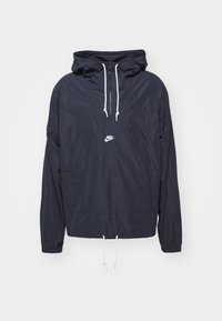 Veste coupe-vent à capuche noire Nike avec cordons blancs, demi fermeture éclair, poche avant et poignets élastiques, présentée sur fond uni.