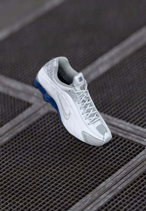 Weißer und grauer Sportschuh mit blauen Details an der Sohle schwebt über einer dunklen Metallgitterfläche.
