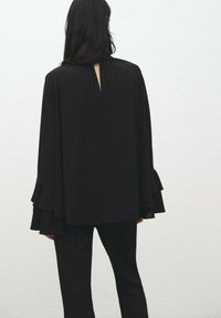 Top negro estilo capa con mangas fluidas y detalle de abertura en la espalda, combinado con pantalones negros ajustados. Textura suave, diseño minimalista.