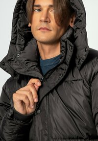 Giacca a vento nera con colletto alto e cappuccio. Presenta una chiusura frontale con zip, bottoni a pressione, polsini in maglia a coste e una superficie liscia e lucida.