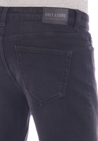 Jean en denim noir avec une poche arrière, une étiquette de ceinture visible et une texture lisse. Le tissu semble léger et durable.