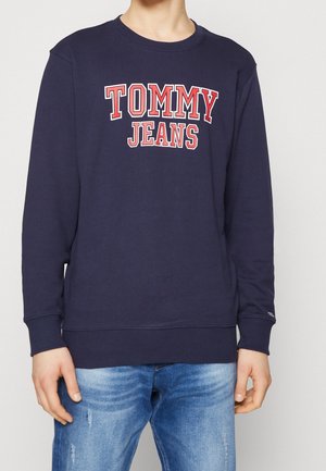 Person trägt einen dunkelblauen Sweatshirt mit „TOMMY JEANS“ in roten und weißen Buchstaben über der Brust, kombiniert mit hellblauen Jeans.