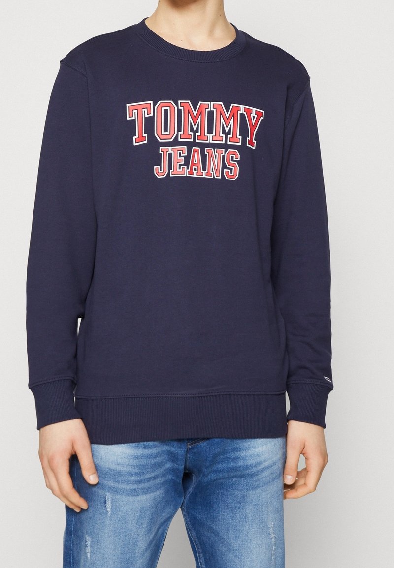Personne portant un sweat-shirt bleu marine avec "TOMMY JEANS" en lettres rouges et blanches sur la poitrine, associé à un jean bleu clair.