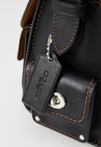 Coin de sac en cuir noir avec des coutures contrastantes orange, un fermoir à torsion argenté et une étiquette de marque Coach suspendue sur une chaîne en métal.