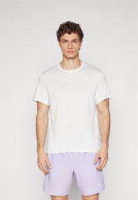 Fehér Nike rövid ujjú póló texturált anyagból, kerek nyakkivágással és diszkrét logóval. Világoslila rövidnadrággal párosítva.