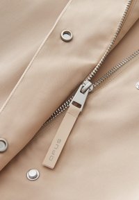 Tessuto beige con una superficie liscia, presenta una cerniera argentata e chiusure a scattare, include un'etichetta con il marchio "OPUS".
