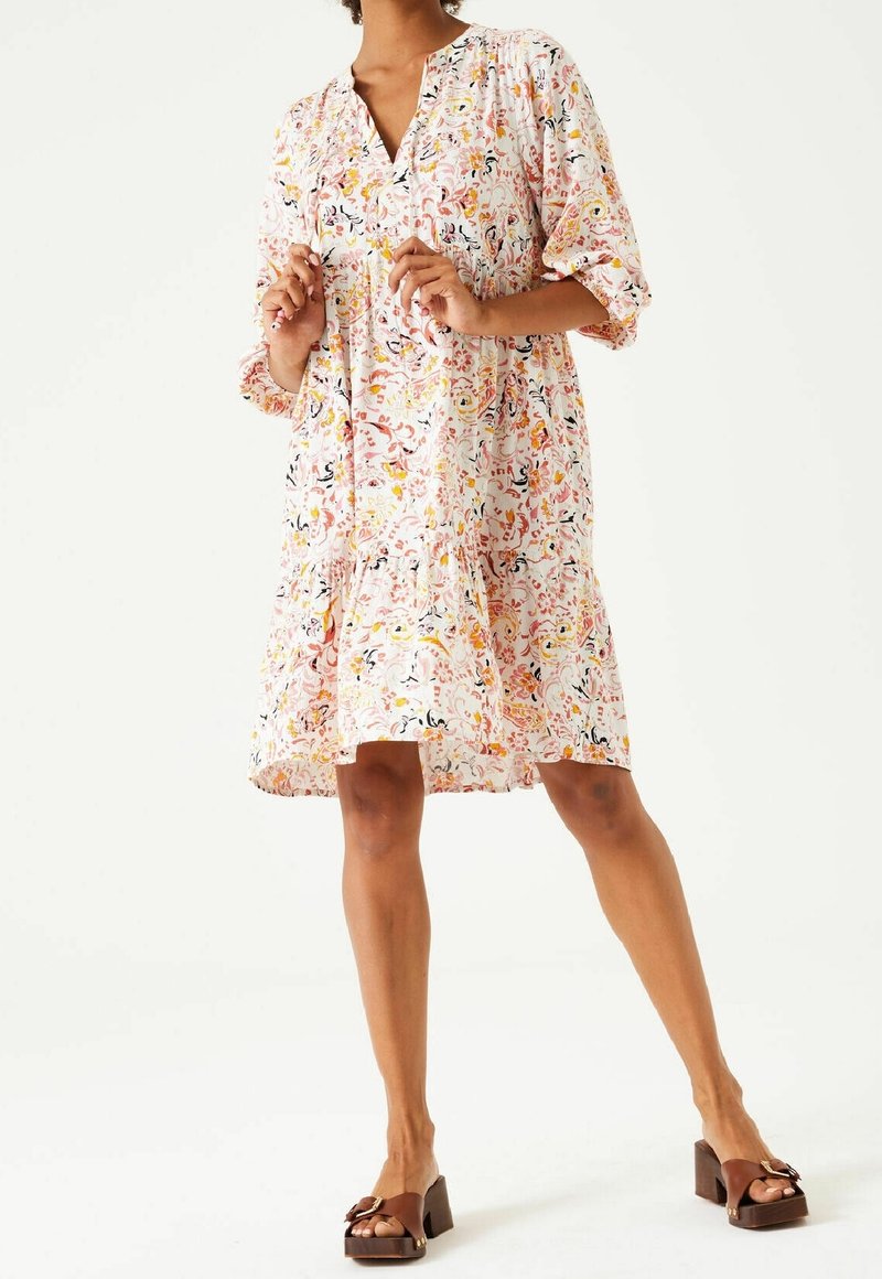 Kobieta w luźnej, kwiecistej sukience do kolan z rękawami 3/4 oraz brązowych sandałach na obcasie, stojąca na tle białego tła.