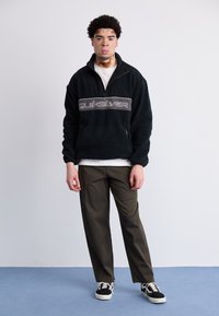 Czarny polarowy sweter z pół zamkiem i beżowym logo "Quiksilver" na piersi. Połączony z oliwkowymi spodniami i czarnymi sportowymi butami.