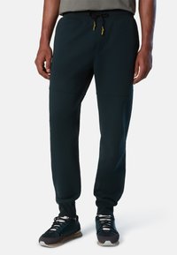 Pantalones jogger ajustados en verde oscuro, fabricados con un tejido suave, con cintura elástica, cordón ajustable y bolsillos. Combinados con zapatillas negras y grises.