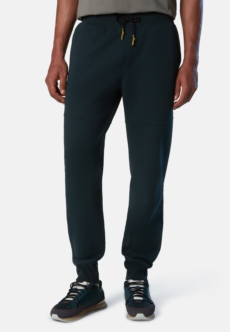 Pantalones jogger ajustados en verde oscuro, fabricados con un tejido suave, con cintura elástica, cordón ajustable y bolsillos. Combinados con zapatillas negras y grises.
