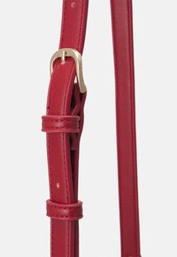 Anse de sac à main en cuir rouge avec une boucle en métal doré et des détails cousus. L'anse est réglable grâce à des nœuds ajustables et présente une texture lisse.