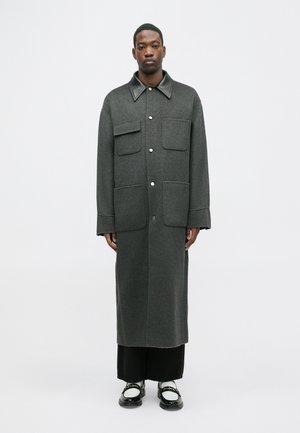 Grauer Wollmantel mit einem schwarzen Lederkragen, zwei Fronttaschen und einer langen Silhouette, kombiniert mit schwarzen Hosen und Schuhen.