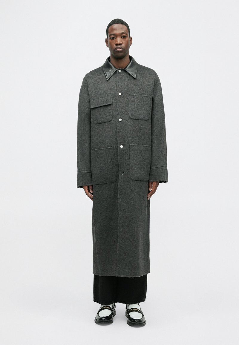 Γκρι μάλλινο παλτό με μαύρη δερμάτινη κολάρα, δύο μπροστινές τσέπες και μακρύ σιλουέτα, συνδυασμένο με μαύρο παντελόνι και παπούτσια.