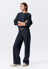 Jean large bleu foncé associé à un sweat-shirt noir oversize ; tissu texturé, design simple, et poignets élastiques. Baskets de couleur claire.