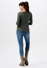 Olijfgroene lange mouwen top, blauwe skinny jeans en beige enkellaarsjes met gestructureerde zolen. Model is van achteren weergegeven.