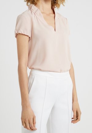 Femme portant un chemisier rose clair à manches courtes avec un col en V et un pantalon blanc taille haute sur un fond uni.