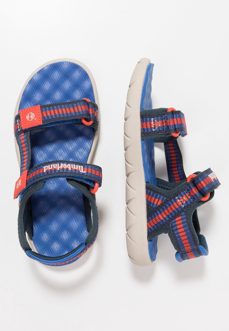 Blå och grå sandaler med justerbara remmar, röda detaljer, texturerad fotbädd och en flexibel beige sula som är lämplig för utomhusbruk.