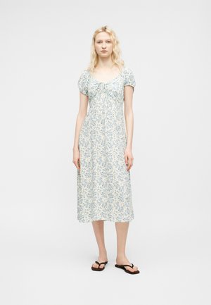 Femme aux cheveux blonds portant une robe midi blanche à imprimé floral bleu, manches courtes bouffantes, et sandales tongs noires, debout devant un fond uni.
