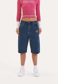 Denim bermuda shorts in dark blue, featuring a front logo embroidery. Styled with a cropped pink knit top and white sneakers.