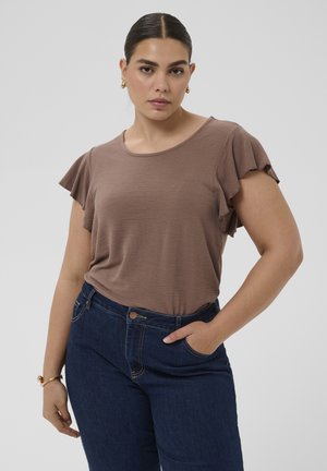 Frau mit dunklen Haaren in einem braunen Kurzarmshirt mit Rüschenärmeln und dunkelblauen Jeans, die mit einer Hand in der Tasche posiert.