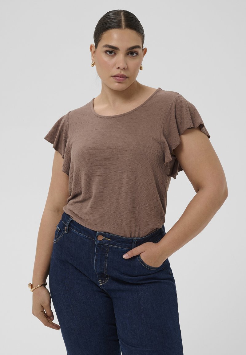 Femme aux cheveux foncés portant un haut marron à manches courtes avec des manches à volants et un jean bleu foncé, posant avec une main dans la poche.