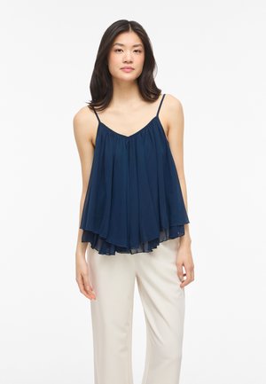 Žena oblečená v tmavomodrom vzdušnom tielku bez rukávov s tenkými ramienkami a krémových širokých nohaviciach, stojaca pred bielym pozadím.