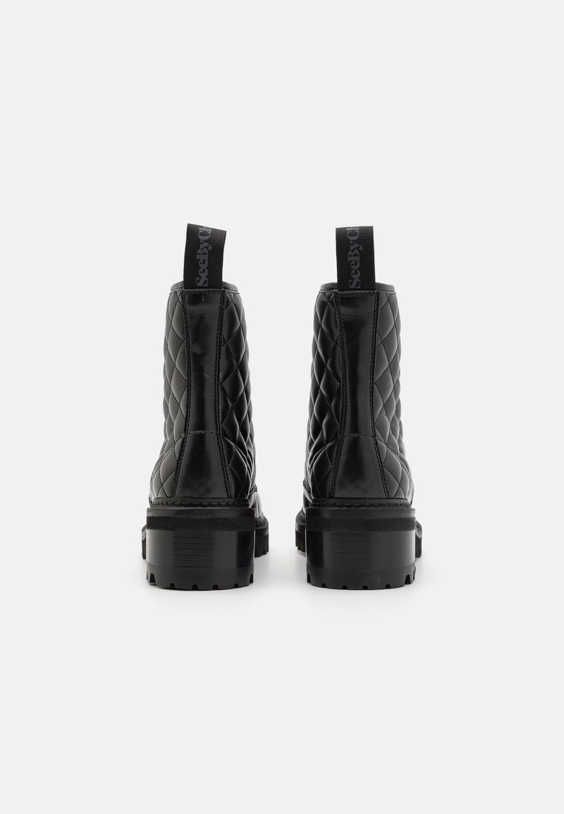 Un paio di stivaletti neri in pelle trapuntata con tacchi spessi, visti da dietro con linguette brandizzate su uno sfondo bianco.