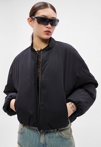 Čierna bomber bunda so zipsom vpredu, oversized strihom a elastickými manžetami, kombinovaná s tmavými slnečnými okuliarmi a vzorovaným topom pod ňou.