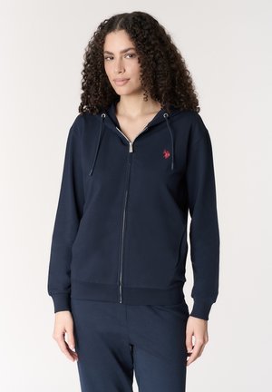 Donna con capelli ricci che indossa una felpa con cappuccio blu navy con zip e un piccolo logo ricamato rosso sul petto, abbinata a pantaloni blu navy coordinati.