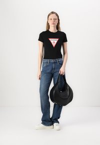 T-shirt noir ajusté avec logo triangulaire rouge 'GUESS?', associé à un jean en denim bleu et un sac à main rond noir. Des baskets blanches complètent la tenue.