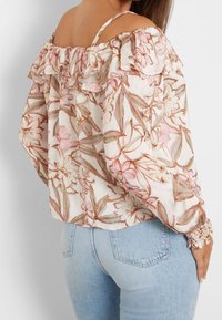 Donna che indossa una blusa floreale con spalle scoperte, rifinita con volant, e jeans in denim leggeri a vita alta, mostrata da dietro e di lato.