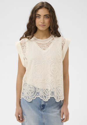 Fiatal nő hosszú, göndör hajjal, ujjatlan krémszínű csipkefelsőt visel egyszínű krémszínű ing felett, világoskék farmernadrággal párosítva.