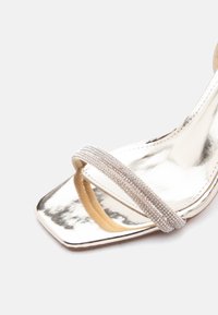 Ασημένιο σανδάλι με ψηλό τακούνι, τετράγωνη μύτη και ένα μόνο λουράκι με λαμπερούς στρας μπροστά.