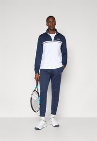 Marineblauer und weißer Trainingsanzug aus leichtem Material, bestehend aus einer Zip-Jacke mit Logo und passenden Hosen, kombiniert mit weißen Sportschuhen und einem Schläger.