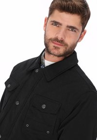 Zwarte jas met een knoopsluiting aan de voorkant, twee borstzakken en een kraag. De textuur heeft een subtiel patroon; gemaakt van duurzaam materiaal.