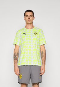 Camiseta deportiva de manga corta en verde claro con patrones geométricos, con un logo de Puma y un emblema del BVB, combinada con pantalones cortos grises.