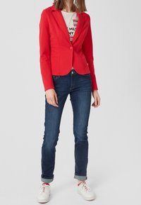 Punainen, istuva bleiseri, jossa on nappikiinnitys, yhdessä tummansiniset farkut. Valkoinen graafinen t-paita sen alla, viimeistelty valkoisilla lenkkareilla.