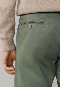 Groene stoffen broek met een gladde textuur, voorzien van één achterzak en een knoopsluiting. De outfit is gecombineerd met een beige gebreide trui.