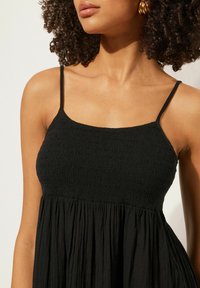 Schwarzes Kleid mit dünnen Trägern; verfügt über strukturierte Raffung am Oberteil und fließende Falten am Rock. Weicher, leichter Stoff.