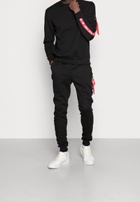Completo felpa nera e pantaloni della tuta in cotone. Presenta un branding rosso sulle maniche e sui lati dei pantaloni, con tasche con zip e sneakers bianche.