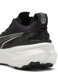 Chaussure de sport noire avec un dessus texturé, un design superposé, des détails réfléchissants et une épaisse semelle blanche avec des accents noirs.
