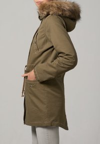 Olivgrön parkajacka med huva foder av fuskpäls, tryckknapppresentation, dragsko i midjan och framfickor. Tyget verkar ha en strukturerad och hållbar kvalitet.