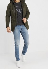 Olive green hooded jacket over a black graphic t-shirt, paired with light blue denim jeans and white sneakers. Casual fit and style.