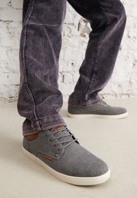 Sneakers di tela grigi con una trama liscia, lacci e dettagli marroni, abbinati a pantaloni scuri a coste su una superficie chiara.